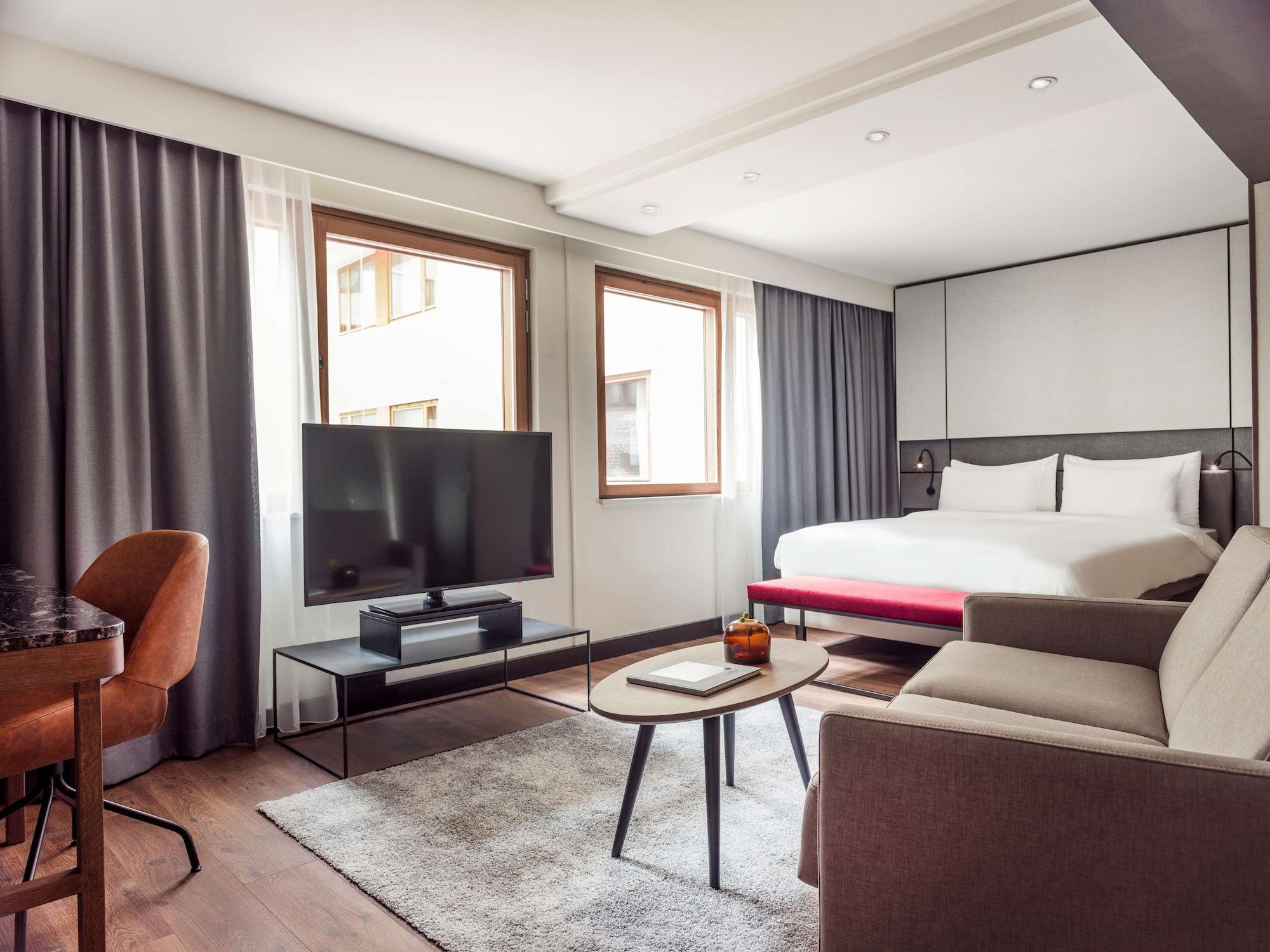 Radisson Blu Royal Viking Hotel, Stoccolma Esterno foto The photo shows a modern hotel room featuring a cozy layout. It includes a large bed with white linens, a comfortable sofa, and a coffee table in the center. There's a flat-screen TV mounted on a black media console. Natural light floods the space th